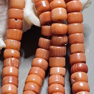 Op de afbeelding: Close-up van een koraal ketting met cilindrische kralen in tinten oranje en perzik. De kralen zijn aan elkaar geregen, waarbij hun natuurlijke textuur en vorm worden getoond. De ketting wordt gepresenteerd tegen een neutrale achtergrond, waarbij de kleur en details van het koraal worden benadrukt.