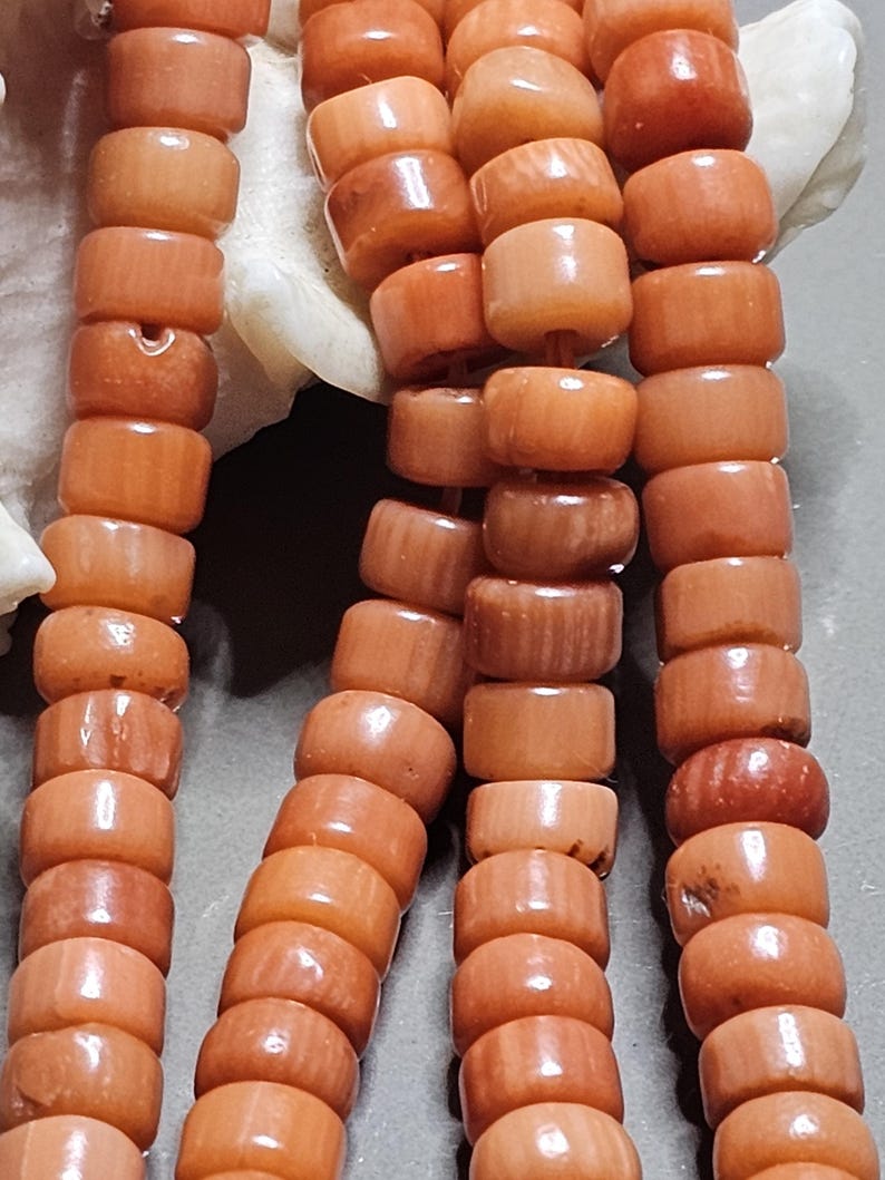 Op de afbeelding: Close-up van een koraal ketting met cilindrische kralen in tinten oranje en perzik. De kralen zijn aan elkaar geregen, waarbij hun natuurlijke textuur en vorm worden getoond. De ketting wordt gepresenteerd tegen een neutrale achtergrond, waarbij de kleur en details van het koraal worden benadrukt.