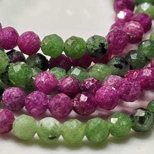 Könnte beinhalten: Ein Strang aus facettierten runden Perlen in leuchtendem Magenta, Dunkelrosa und verschiedenen Grüntönen. Die Perlen haben eine texturierte, mehrfach facettierte Oberfläche, die das Licht reflektiert, wobei einige grüne Perlen kleine schwarze Einschlüsse aufweisen.