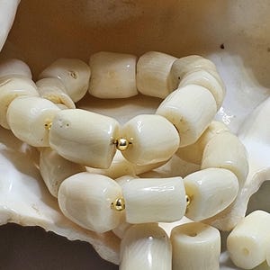 May include: Cream-colored bracelet made of large, cylindrical beads. The beads are strung together with gold-colored accents. The bracelet is displayed on a white seashell.