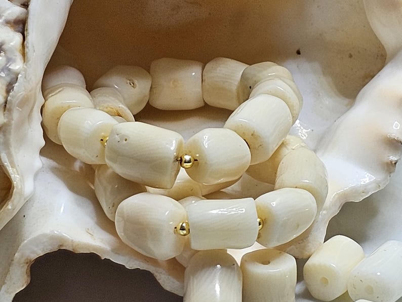 May include: Cream-colored bracelet made of large, cylindrical beads. The beads are strung together with gold-colored accents. The bracelet is displayed on a white seashell.