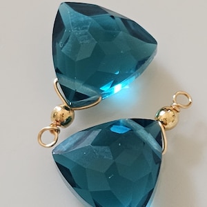 Op de afbeelding: Twee turquoise, driehoekige edelsteenhangers met goudkleurige accenten. Elke hanger heeft een facetgeslepen oppervlak en is verbonden met een goudkleurige kraal en een kleine lus. De hangers worden op een wit oppervlak gepresenteerd.