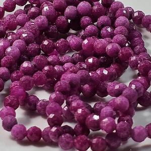 Könnte beinhalten: Nahaufnahme von mehreren Strängen facettierter, runder Perlen in einem tiefen Magenta. Die Perlen haben eine glitzernde, strukturierte Oberfläche und sind aufgefädelt. Das Bild zeigt mehrere Stränge, was auf eine Vorrat an Perlen für die Schmuckherstellung hindeutet.