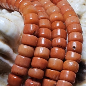Op de afbeelding: Close-up van strengen koraalkralen. De kralen hebben een warme, rood-oranje kleur en zijn cilindrisch van vorm. De kralen zijn aan elkaar geregen, waardoor een ketting of armband ontstaat. De achtergrond is licht en neutraal.
