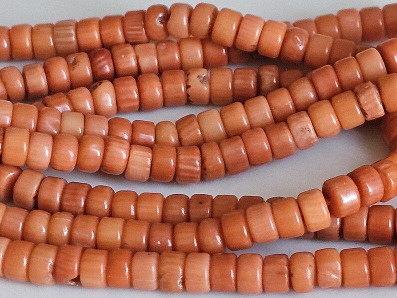Op de afbeelding: Close-up van strengen koraalkralen. De kralen zijn cilindrisch, met een warme oranje tint. De kralen zijn aan elkaar geregen, waardoor een textuur en organisch uiterlijk ontstaat. De kralen worden waarschijnlijk gebruikt voor het maken van sieraden.