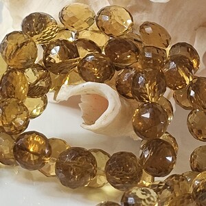 May include: A close-up of a strand of faceted, golden-brown gemstone beads. The beads are round with multiple facets, reflecting light. The beads are resting on a white surface with a seashell in the background. The beads are a warm, amber color.