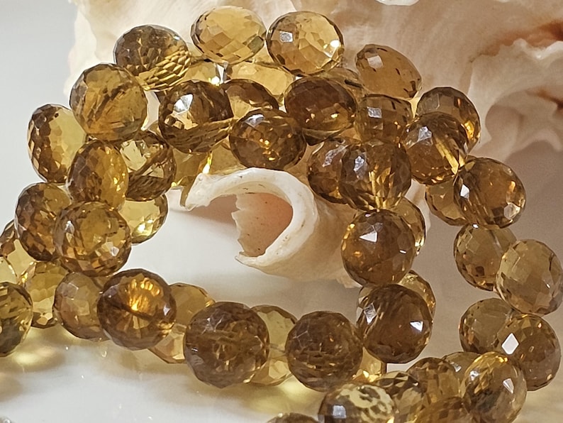 May include: A close-up of a strand of faceted, golden-brown gemstone beads. The beads are round with multiple facets, reflecting light. The beads are resting on a white surface with a seashell in the background. The beads are a warm, amber color.