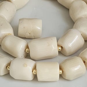May include: Close-up of a necklace featuring large, off-white, barrel-shaped beads. The beads are strung together with gold-colored spacers. The beads have a natural, textured appearance, and the necklace is displayed on a light-colored surface.