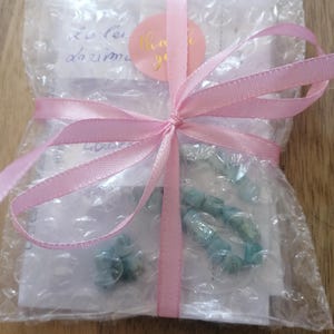 May include: A clear bubble wrap pouch containing light blue stone beads, secured with a pink ribbon tied in a bow. A round pink sticker with the gold text thank you is placed on the front. White paper inserts are visible inside the packaging.