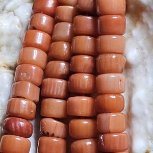 Op de afbeelding: Close-up van een koraal kralenketting. De kralen hebben een warme, rood-oranje kleur en zijn cilindrisch van vorm. De kralen zijn aan elkaar geregen, waardoor een ketting ontstaat. De achtergrond is licht, gebroken wit.