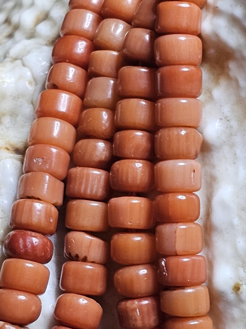 Op de afbeelding: Close-up van een koraal kralenketting. De kralen hebben een warme, rood-oranje kleur en zijn cilindrisch van vorm. De kralen zijn aan elkaar geregen, waardoor een ketting ontstaat. De achtergrond is licht, gebroken wit.