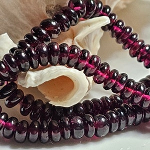 Puede incluir: Una hebra de cuentas de vidrio de color morado oscuro, cada cuenta es redonda y ligeramente aplanada, creando un aspecto único y texturizado.