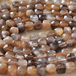 Puede incluir: Un collar de cuentas de piedras preciosas facetadas en tonos marrones, beige y grises. Las cuentas tienen forma geométrica y están ensartadas en un hilo naranja. Adecuado para la fabricación de joyas.