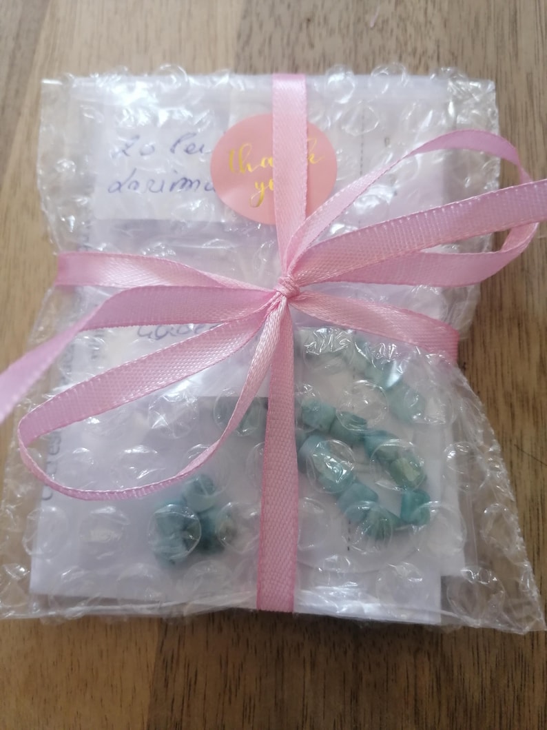 May include: A wrapped package with a light pink ribbon tied in a bow. The package is wrapped in bubble wrap and contains a light blue beaded bracelet. A pink sticker with the words "Thank you" is visible.
