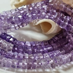 Könnte beinhalten: Nahaufnahme von Strängen aus Amethystperlen. Die Perlen sind eine Mischung aus hellen und dunklen Lilatönen mit einer facettierten, quadratischen Form. Die Perlen sind in einem gebogenen Muster angeordnet, mit einer Muschel im Hintergrund.