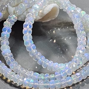 May include: A strand of faceted, translucent white beads with an iridescent, opalescent sheen. Each small, circular bead features multiple flat surfaces that catch the light, creating subtle flashes of blue, green, and pink colour throughout.