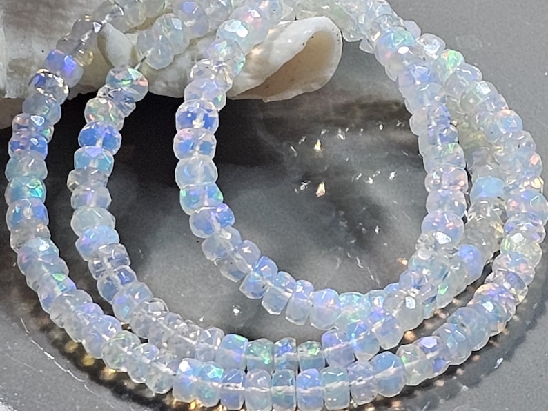May include: A strand of faceted, translucent white beads with an iridescent, opalescent sheen. Each small, circular bead features multiple flat surfaces that catch the light, creating subtle flashes of blue, green, and pink colour throughout.