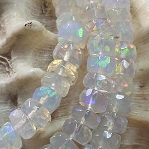 May include: A strand of faceted, translucent white beads with an iridescent, opalescent sheen that reflects flashes of green, blue, and yellow light. The beads are circular with a flat profile and are strung together on a thin, light-coloured cord.