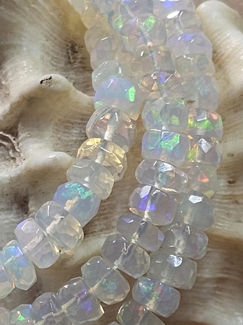 May include: A strand of faceted, translucent white beads with an iridescent, opalescent sheen that reflects flashes of green, blue, and yellow light. The beads are circular with a flat profile and are strung together on a thin, light-coloured cord.