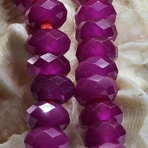 Könnte beinhalten: Zwei parallele Stränge aus facettierten, durchscheinenden magentafarbenen Perlen, die auf eine leuchtend orangefarbene Schnur aufgezogen sind. Jede Perle hat ein kreisförmiges, facettiertes Design, das das Licht reflektiert und ein Muster erzeugt.