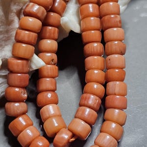 Op de afbeelding: Close-up van een koraal ketting met strengen van cilindrische, oranje kralen. De kralen zijn aan elkaar geregen, wat een natuurlijke, organische uitstraling geeft. De ketting wordt gepresenteerd tegen een neutrale achtergrond, waardoor de kleur en textuur van het koraal worden benadrukt.