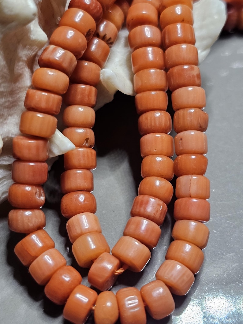 Op de afbeelding: Close-up van een koraal ketting met strengen van cilindrische, oranje kralen. De kralen zijn aan elkaar geregen, wat een natuurlijke, organische uitstraling geeft. De ketting wordt gepresenteerd tegen een neutrale achtergrond, waardoor de kleur en textuur van het koraal worden benadrukt.