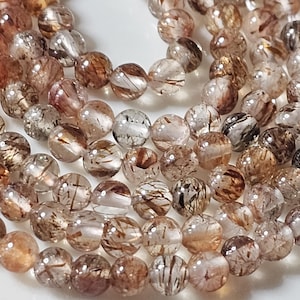 Può includere: Primo piano di una collezione di perline rotonde e trasparenti con inclusioni di minerali marroni e neri. Le perline sono infilate insieme, creando un effetto a strati. Le perline hanno un aspetto lucido e brillante.