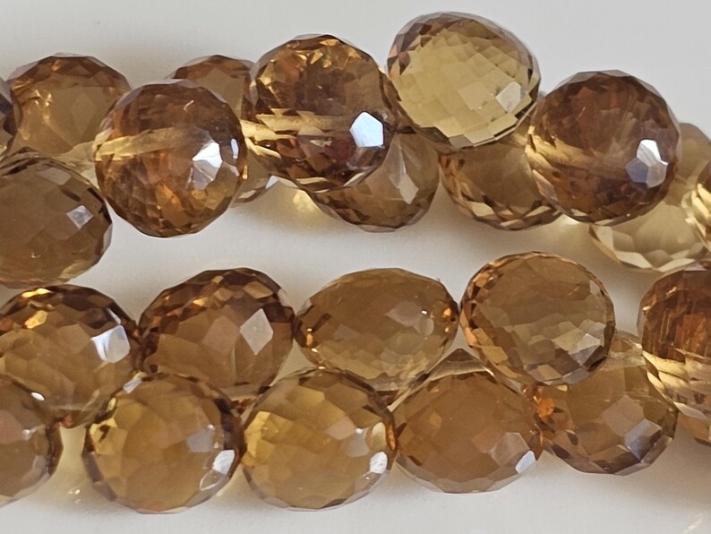May include: Close-up of faceted, round, amber-colored gemstone beads. The beads have a polished, reflective surface and are strung together. The gemstones are a warm, earthy tone, and the facets catch the light.