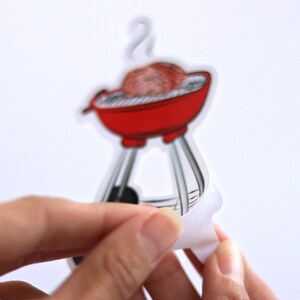 May include: A red and black grill with a single hamburger patty on the grill. The grill is on a white background and has a white backing paper peeling off.