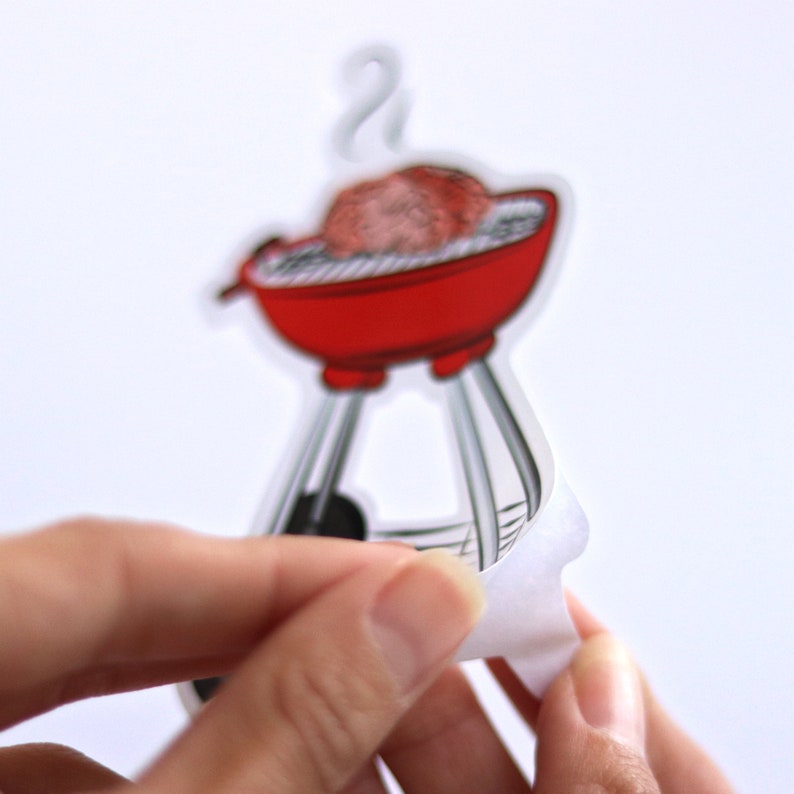 May include: A red and black grill with a single hamburger patty on the grill. The grill is on a white background and has a white backing paper peeling off.