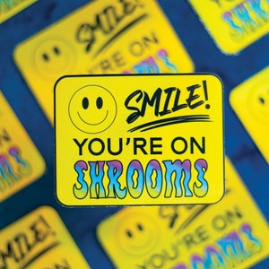 Peut inclure: Autocollant jaune avec un smiley et le texte "Smile! You're on Shrooms" en noir et lettres arc-en-ciel.