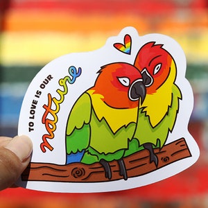May include: A sticker featuring two lovebirds perched on a branch. The birds are green, yellow, orange, and red. The sticker reads "To love is our nature" in rainbow lettering. A rainbow heart is above the birds.