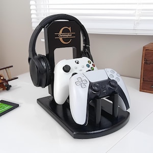 May include: Black wooden gaming headset and controller stand with the name "Christopher" and a "C" initial. The stand holds a black headset and two white game controllers. A small wooden airplane and a small wooden cabinet are in the background.