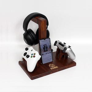 Op de afbeelding: Een houten gaming-console standaard met een zwarte headset, een witte controller, een zilveren controller en een smartphone. De standaard is gegraveerd met het woord "RONIN".