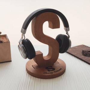 May include: Wooden headphone stand shaped like the letter "S" with the name "Tracey Salazar" engraved on the base. The stand is made of light brown wood and has a dark brown finish. A pair of black headphones are resting on the stand.