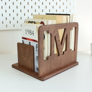 May include: A dark brown wooden book stand with the letter "M" cut out of the front. The stand is holding a stack of books, including "1984" by George Orwell.