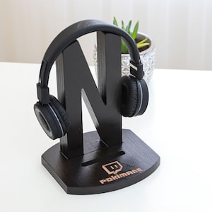 Op de afbeelding: Zwarte houten koptelefoonstandaard in de vorm van de letter N met een zwarte koptelefoon erop. De standaard heeft de tekst "Pokimane" en een Twitch-logo gegraveerd op de basis.