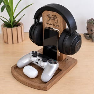 May include: A wooden gaming organizer stand featuring a vertical support with a laser-engraved game controller icon and the text RAPTOR. The stand holds black over-ear headphones, a smartphone, a silver game controller, and a white earbud case.