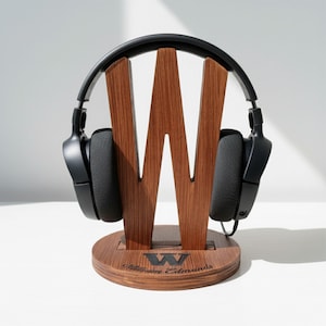 May include: A wooden headphone stand with a large "W" design, holding a pair of black headphones. The stand is made of dark wood and has a circular base with the name "William Edmunds" engraved on it. The headphones are black.