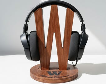 Pode incluir: Um suporte de madeira para fones de ouvido com um design de "W" grande, segurando um par de fones de ouvido pretos. O suporte é feito de madeira escura e tem uma base circular com o nome "William Edmunds" gravado. Os fones são pretos.