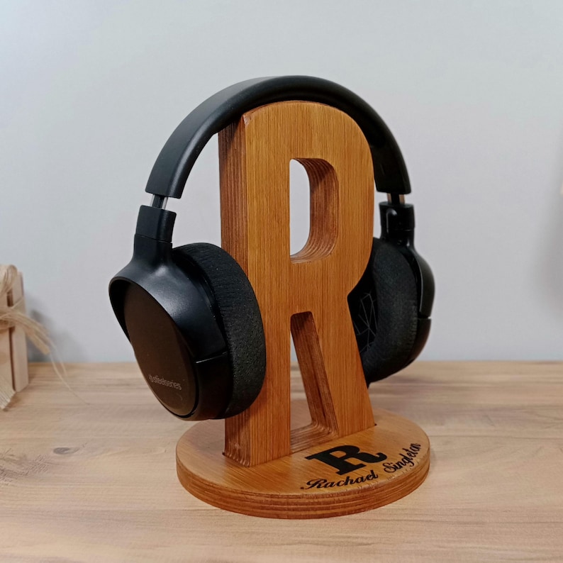 May include: A wooden headphone stand shaped like the letter "R" with a pair of black headphones resting on it. The stand is made of light brown wood and has a circular base. The name "Rachael Singleton" is engraved on the base.