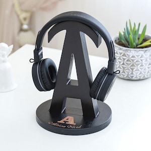 May include: Black wooden headphone stand shaped like the letter "A", with a pair of black headphones resting on it. The base of the stand has the name "Adrian Ward" and the letter "A" in a copper color.