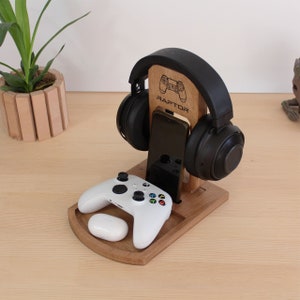 Op de afbeelding: Houten standaard met een telefoonhouder, een koptelefoonhouder en een controllerhouder. De standaard is gegraveerd met het woord "RAPTOR" en een videogamecontrollerpictogram.