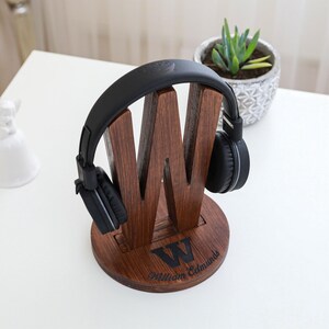 May include: A wooden headphone stand in the shape of the letter "W" with a pair of black headphones resting on it. The stand is engraved with the letter "W" and the name "William Edmunds". A small potted plant is in the background.