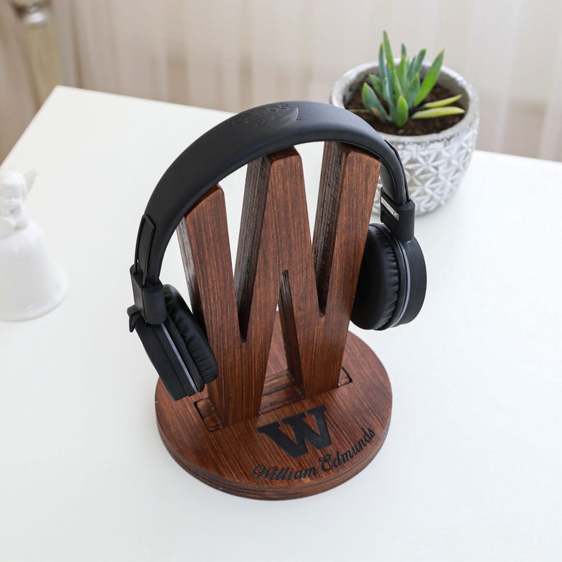 May include: A wooden headphone stand in the shape of the letter "W" with a pair of black headphones resting on it. The stand is engraved with the letter "W" and the name "William Edmunds". A small potted plant is in the background.