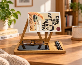 Puede incluir: Un organizador de escritorio de madera con un soporte inclinado que sostiene un libro titulado Cengiz Aytmatov Toprak Ana, una bandeja para un smartphone, unas gafas de montura negra y una ranura con dos bolígrafos negros.