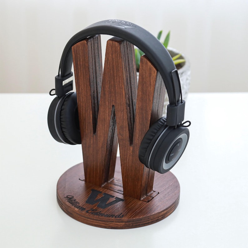 May include: A wooden headphone stand in a dark brown finish, shaped like the letter "W". The stand holds a pair of black headphones with silver accents. The base of the stand is round and has the name "William Edmunds" engraved on it.