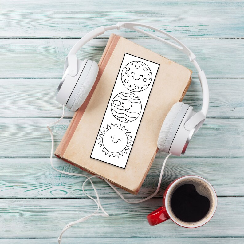 May include: A white book with a black and white bookmark featuring a sun, a planet, and a star with smiling faces. The book is on a blue wooden surface with a pair of white headphones and a red coffee mug.