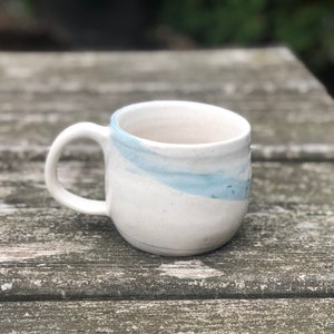 May include: A white ceramic mug with a blue glaze design. The mug has a rounded shape and a handle.