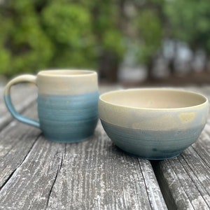May include: A blue and cream ceramic mug and bowl with a subtle ombre effect. The mug has a handle and the bowl is round. Both are sitting on a wooden surface.